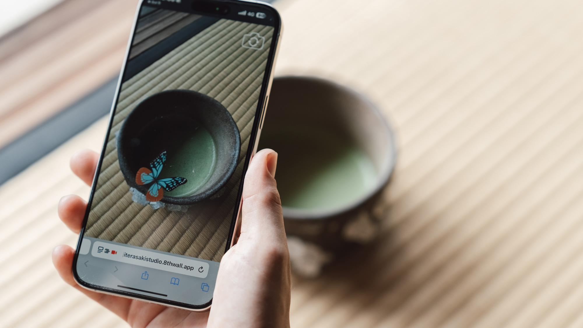 A hand holds a smartphone displaying a photo of a matcha bowl with a digital butterfly overlay. The real bowl, filled with green tea, sits on a tatami mat.
